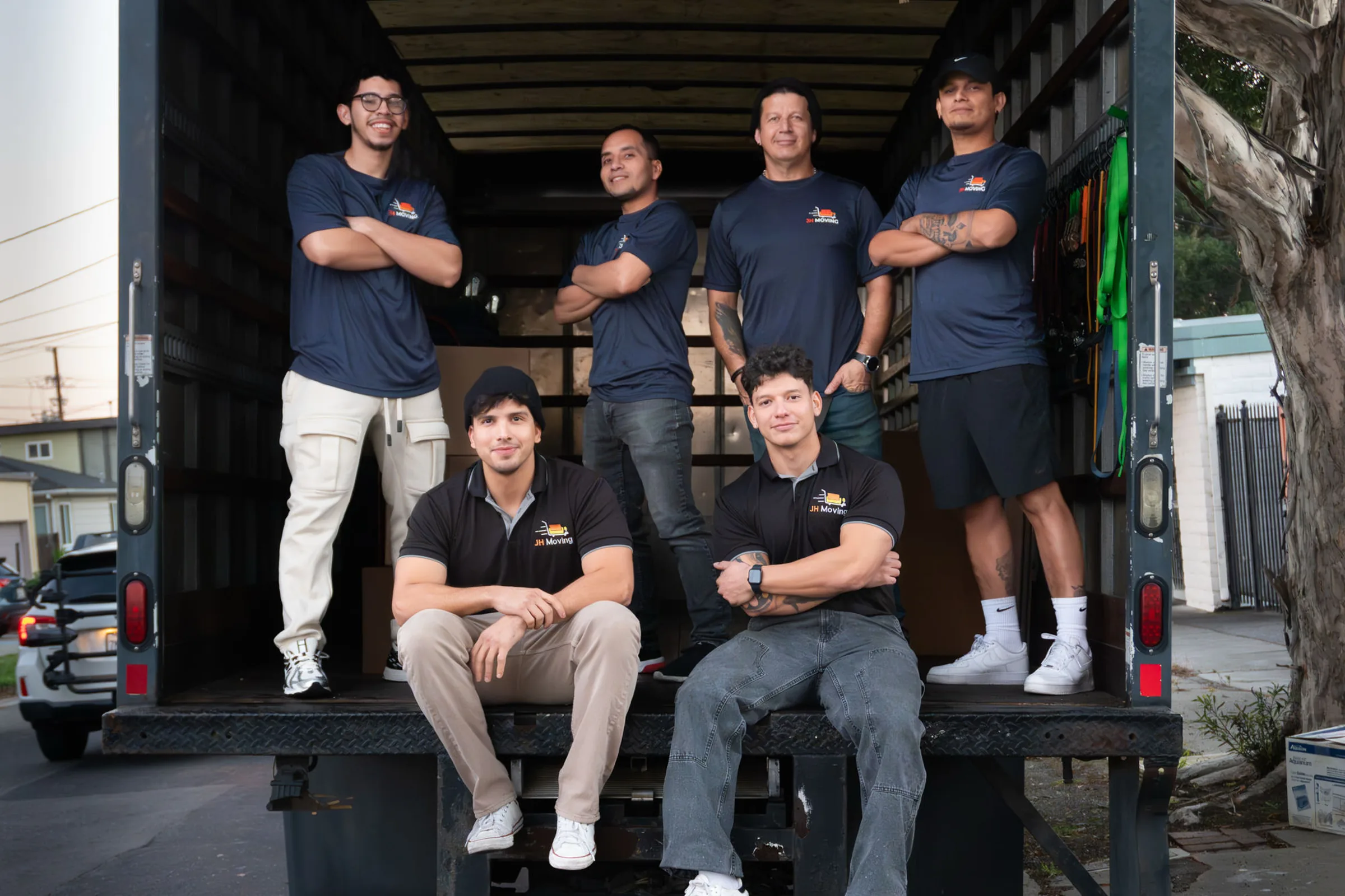 The JH Moving team, six people in company shirts, sitting and standing in the open back of their truck at the end of a day.
